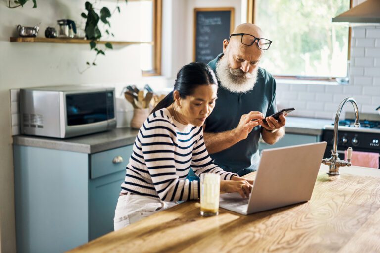 Woman and man in a kitchen standing over a computer. Man is holding a device.