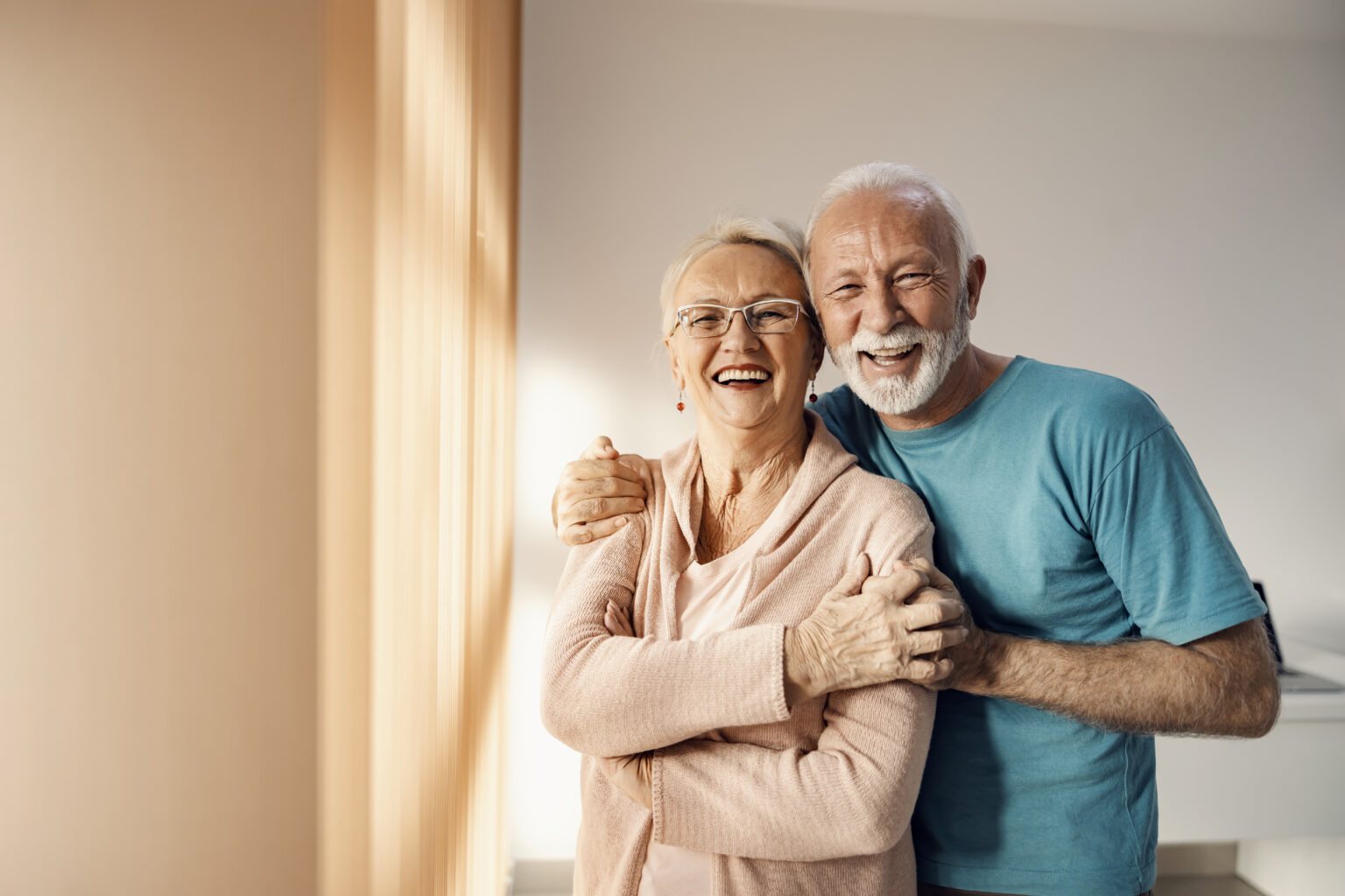 Senior couple hugging in a nursing home. A happy senior couple standing next to a window in a nursing home, hugging and smiling. They have all care they need.