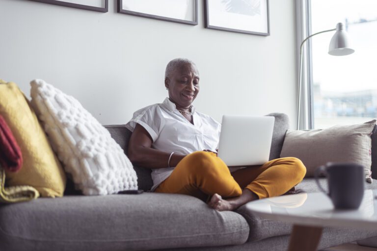 A black senior woman sits comfortably on her couch in her apartment, typing on her laptop.