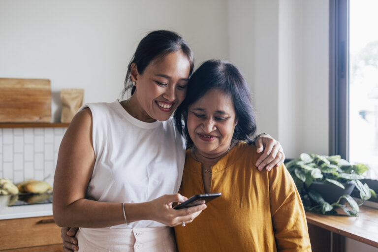 A smiling Asian female hugging her mother while they are reading something online using their black smartphone.