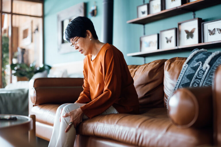 A senior Asian woman suffering from pain in leg while sitting on sofa in the living room at home