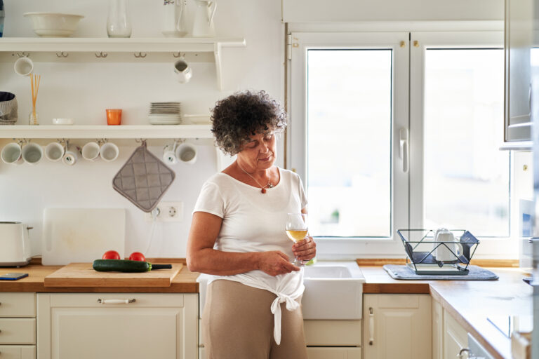 Front view of a relaxed mature woman holding a glass of wine looking down in kitchen.